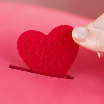 Piggy bank. female hand puts Red heart into slot of red donation box. Concept of sincere devotion to faith. Concept of donorship, life saving or charity. Valentines Day. Close-up shot