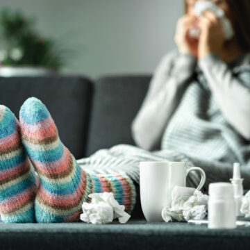 Sick woman with flu, cold, fever and cough sitting on couch at home. Ill person blowing nose and sneezing with tissue and handkerchief. Woolen socks and medicine. Infection in winter. Resting on sofa.